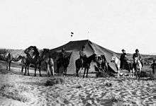 Photograph of desert tent, mounted soldiers and camels