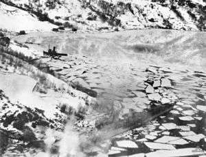 Two ships close to the shore of a body of water near steep snow-covered hills. Much of the body of water is covered by sheets of ice.