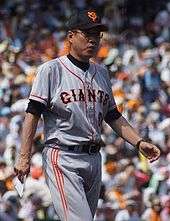 Japanese man wearing a gray, black and orange uniform walks in front of a blurry crowd