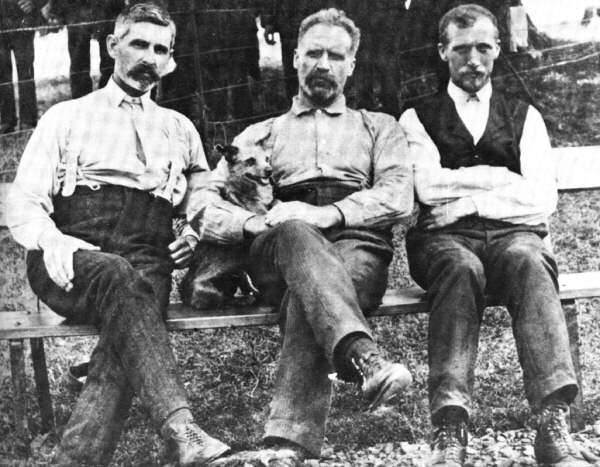 Old photograph depicting three bearded men seated outdoors on a bench with the man in the center holding a small dog