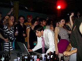 A Caucasian man wearing a tie performs music using a laptop. Several people dance around him.