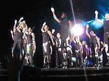 Left profile of a group of people jumping onstage with their right hand stretched up. They are wearing black tops and black-and-white striped pants and wrist bands. They have white gloves on their right hand. They are illuminated by white light from above.