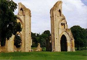 Two ruin parts of walls with arched doorways, surrounded by grass and trees.