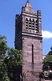 A church tower in a robust style with a short stone spire and four louvred openings in the bell chamber near the top.