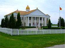 A photograph of a two-story building. The roof is topped with a large cupola and three visible kalashes. Golden letters on the pediment say "CAPITAL OF THE GLOBAL COUNTRY OF WORLD PEACE". There is a circular driveway with a flagpole bearing the GCWP's sunburst flag. The building and drive are surrounded by a white picket fence, green lawn, and shrubbery.