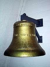 A large brass bell, engraved with the words "S.M.S. Von Der Tann", suspended on a white brick wall.