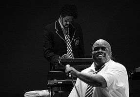 In the forefront, a man in a white dress shirt and tie holding a microphone on a stand. In the background, a man wearing a jacket, tie, and sunglasses playing keyboards.