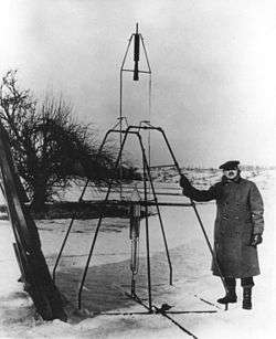 This black and white photo shows Robert Goddard standing next to a frame in which a small (three to four foot) rocket is mounted. It is winter, and open snow-covered fields are in the background.
