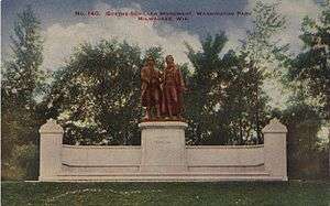 Picture postcard of a large bronze statue of two men. The statue is on top of a stone pedestal that is flanked on each side by stone walls with stone benches running along the base of the wall. The pedestal, walls, and benches sit on top of a large stone step. The monument has grass in front of it, and trees behind it. There is printing along the top of the postcard that says "No. 140. Goethe–Schiller Monument. Washington Park. Milwaukee, Wis."