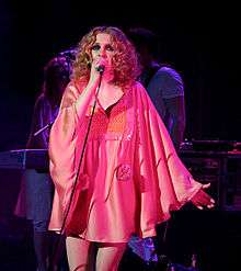 A woman with blonde curly hair wearing a pink dress and singing into a microphone