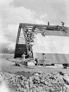 Workmen in slouch hats lay corrugated iron on a large semi-cylinder shaped structure with a metal frame.