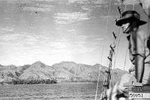 A man in a slouch hat looks out from a sailing ship over a mountainous coastline.