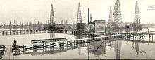 A black-and-white photograph showing a field of oil derricks and thin wooden boardwalks built over the water.