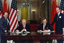 Two men in suits sit signing documents at a large table in front of their country's flags. Two others stand outside watching them.