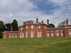 A large brick mansion. The central rectangle of the house has large windows with rounded tops. Wings nearly the width of the central structure extend to either side.