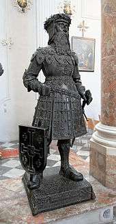 A statue of a knight with a long beard. He is wearing a crown of thorns and elaborate armour. He has a sword in his left hand, and a shield rests against his right leg.