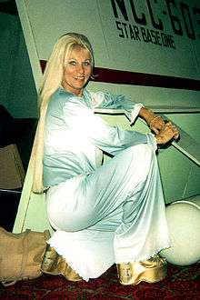 A blonde Caucasian woman kneels, while wearing a light blue flowing 70's style outfit.