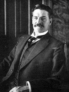 Photograph of a moustached middle-aged man in a dark suit and waistcoat, sitting in a chair while looking at the camera