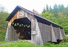 A brown wooden covered bridge with pointed roof. Above its portal are signs saying "Millbrook 1902" and "Pedestrians only". To the right a faded sign says "Safe load 6 tons"