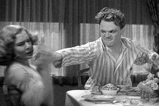 Cagney, in stripped pajamas, looks angry as he reaches across a breakfast table with the grapefruit in his hand.
