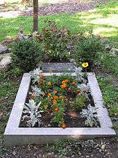 A rectangular stone slab bearing Lad's name and dates of birth and death lies above a rectangular plot of earth lined with concrete. Flowers are planted on the grave and around the slab.
