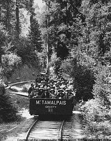 A single open railroad car, on a single rail track that twists and turns through a forest. The car is carrying about 30 passengers, all of whom are wearing hats.