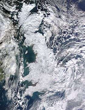 A satellite photo of Great Britain and Ireland. Great Britain is white as a result of snow cover and Ireland is mostly green with some snow cover in the east. Cloud is scatted across the surrounding sea and eastern Ireland, as well as the east coast of Great Britain (especially the southeast).