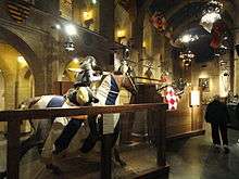  The interior of the Higgins armory. Large stone walls lined with suits of armor and a set of mannequins of men on horses jousting.