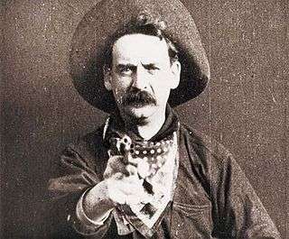 A black-and-white screenshot of a man facing the camera, while also pointing a revolver forward. He has a large mustache and is wearing a cowboy hat, a bandana around his neck, and a heavy jacket.