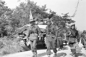 Two men in uniform walking along a road, the man on the left is wearing a slouch hat and holding a map, while the man on the right is wearing a beret. Both men binoculars around their necks. In the background is a stationary tank on the side of the road and a number of armed soldiers.