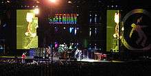 A man wearing a hat is captured on two screens running across a stage as several musicians perform in front of an audience. "GREENDAY" is seen flashed in multi-colored letters on another screen.