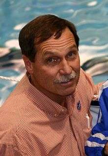 Head and shoulders portrait of Gregg Troy, dark-haired, 58-year-old man with grey mustache, shown in an orange-and white checkered shirt.