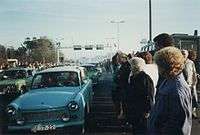 East German Trabant cars driving between dense crowds of people. Metal gantries over the road and a watchtower are visible in the background.
