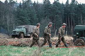 Three uniformed men, armed with assault rifles, walking in a column through a grassy landscape with dense trees in the background. A canvas-sided truck is visible in the left background and part of another vehicle is seen on the right in a stretch of ploughed-up ground.