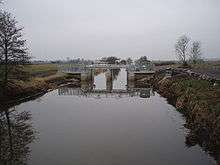 Straight watercourse, surrounded by fields and crossed by metal and concrete structure.