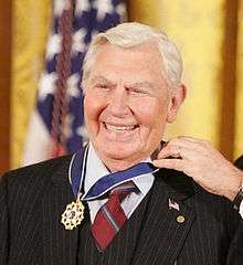 A man with white hair dressed in a black suit, wearing the Presidential Medal of Freedom