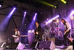 A rock band performing live on-stage. The stage is brightly lit On the left, a man plays bass behind a keyboard. In the centre, a man plays guitar and behind him a man plays drums. On the right, a man plays an electric mandolin.