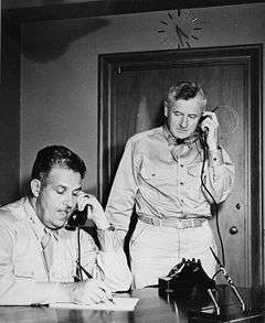 Two men in shirts and a ties. One is sitting at a desk and the other standing. Both are talking on telephones.