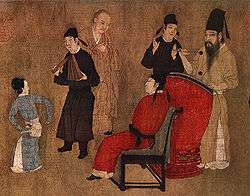 A small section of a larger painting of a party. In the center, a man in red robes is seated in a chair. In front of him is a small female dancer, a male musician dressed in black, and a guest. Behind the chair is a second guest and a man in brown robes hitting a man sized drum with drumsticks.