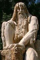  Sculpture of a mature bearded man with long hair locks that cover his nudity. The man stands on his left foot, his right foot and left hand rest atop a wet mossy rock. He has a peaceful facial expression and gazes at the viewer, his left shoulder and arm protrude from under his thick hair locks and show prominent musculature.