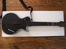 A black guitar controller sits on a white piece of paper and a wooden floor.