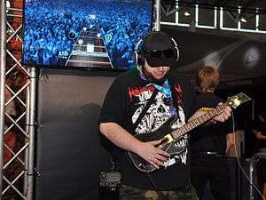 Man wearing headphones plays Guitar Hero Live in a convention. The in-game footage is displayed at a screen behind him.