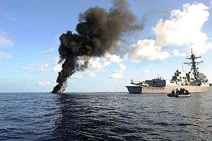 A tall plume of black smoke rises from the blue ocean waters next to a large gray battleship and a small black inflatable boat.