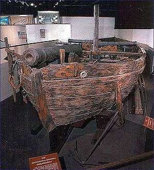 The ship is shown in a museum installation, mounted on a wooden framework. The ship's woodwork has been discolored, and the ship sports a short mast, a single cannon facing forward near the bow, and two cannons facing port and starboard respectively near the stern.