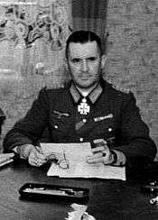 A man wearing a military uniform with an Iron Cross displayed at the front of his uniform collar sits behind a desk.