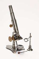 A stand microscope created by Joseph Gutteridge, a weaver from Coventry. The microscope allows coarse and fine adjustments, and its height is adjustable by a set screw. It consists of two objectives each with a screw on cap and a slidecarrier that is moveable laterally. The mirror also has a cap. It has a bench magnifier, lens moveable through 360° and has a base weighted with lead.