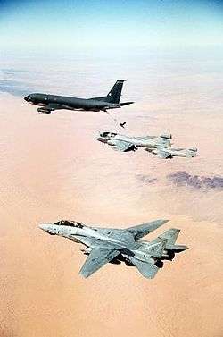 Portrait photography of four aircraft overflying orange desert and almost-flat terrain; horizon is blurred. Leading is black aircraft, followed by two single-engine jet aircraft, the one closer to camera being refueled by leading jet via a stiff hose connecting the two. Closest jet to camera is pale gray, has two engines and vertical fins, flying with wings unswept.