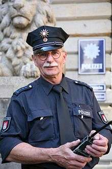 A man wearing a blue shirt with the word "POLIZEI" written on it, Epaulets with 5 stars on them, and a blue peaked cap with a 12-pointed star on it. He is holding a radio.