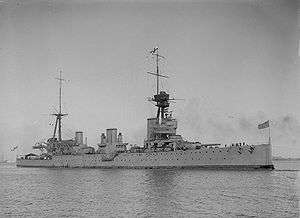 Front oblique view of a large warship at anchor with two masts, three funnels, and two gun turrets visible