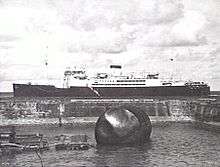 Black and white photo of a large ship anchored off a beach. A damaged buoy is in the foreground of the photo.